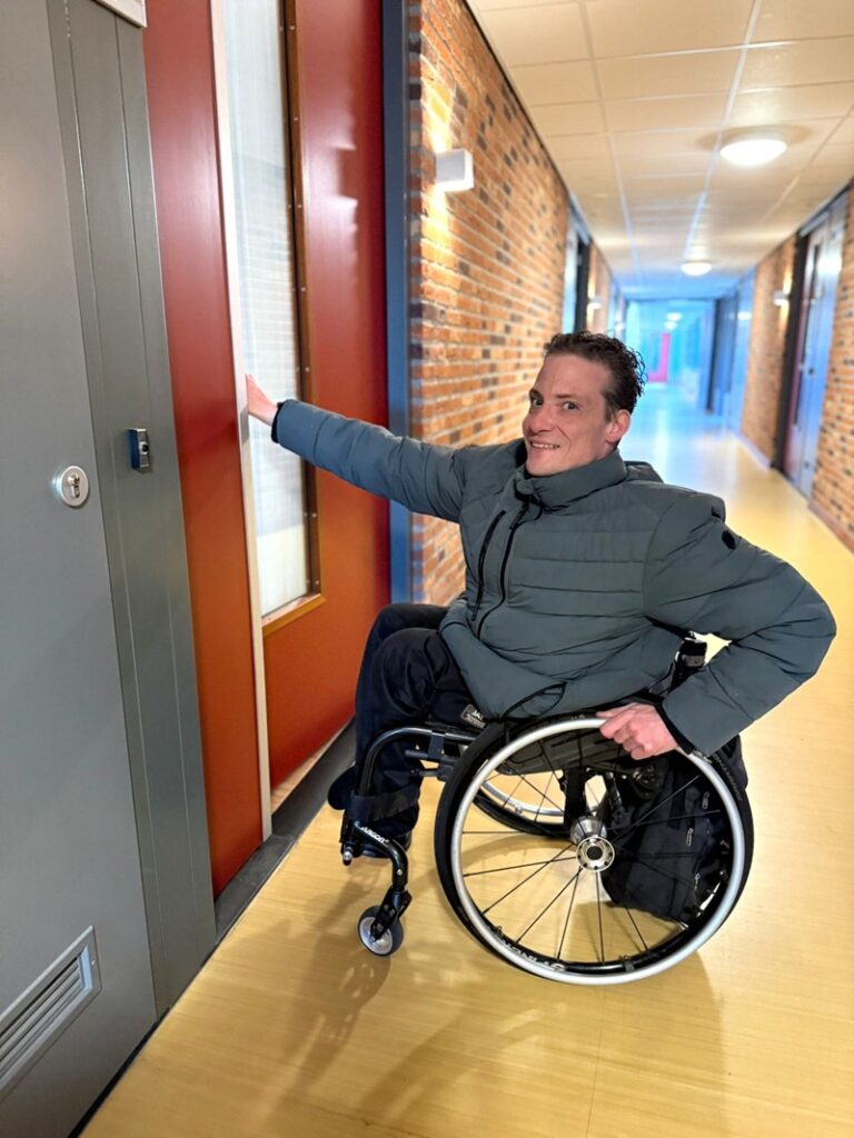 Bewoner Sten Cremers, een jonge man in een rolstoel, voor de deur van zijn appartement.